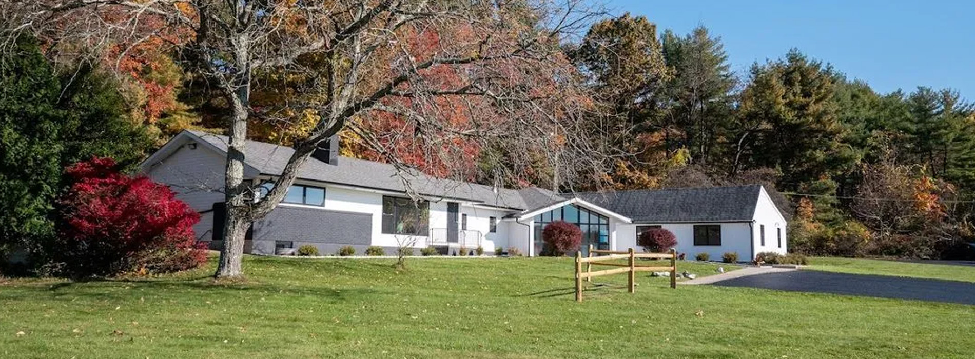The image shows a two-story residential house with a white exterior, situated on a grassy area surrounded by trees with autumn foliage.