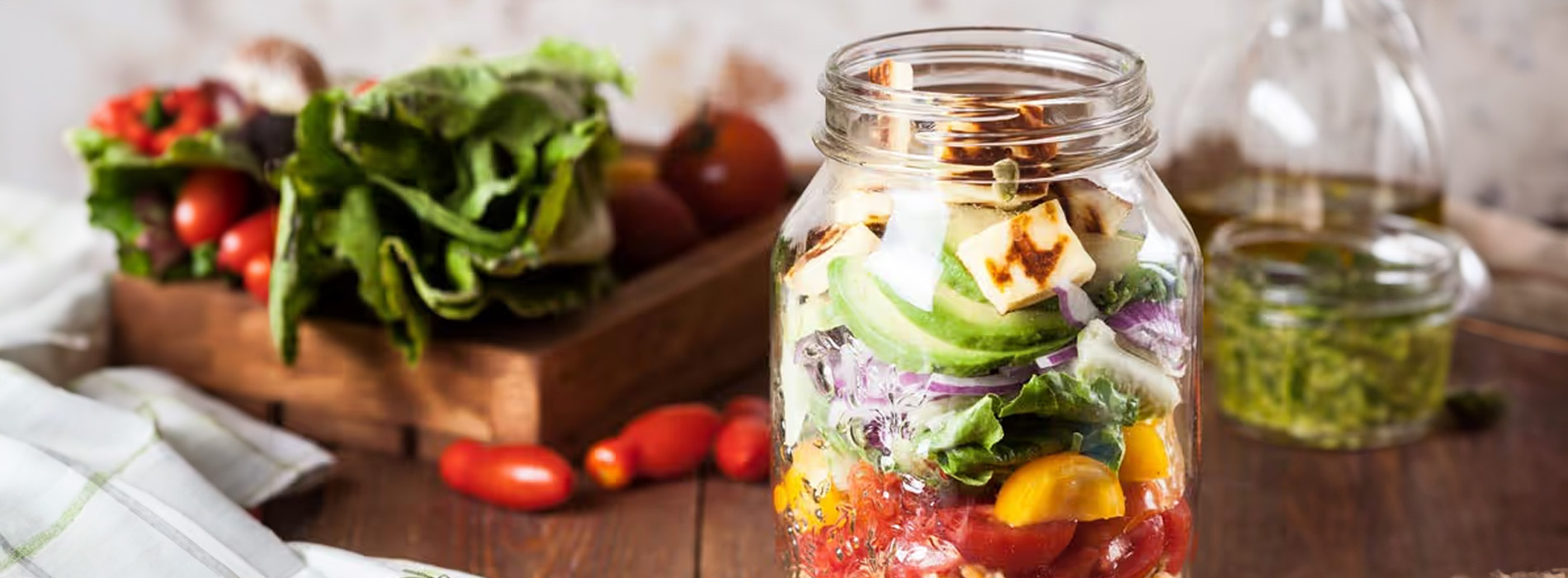 The image shows a jar filled with fresh vegetables such as tomatoes, lettuce, and cheese, placed on a table surrounded by other food items like broccoli and carrots, suggesting a focus on healthy eating and meal preparation.