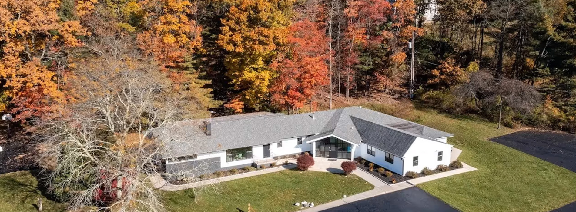 The image shows a two-story house with a white exterior and gray roof, surrounded by trees with autumn foliage, set against a clear sky.