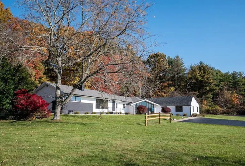The image depicts a two-story house with a garage on the left side, set against a backdrop of autumn trees and a clear sky.