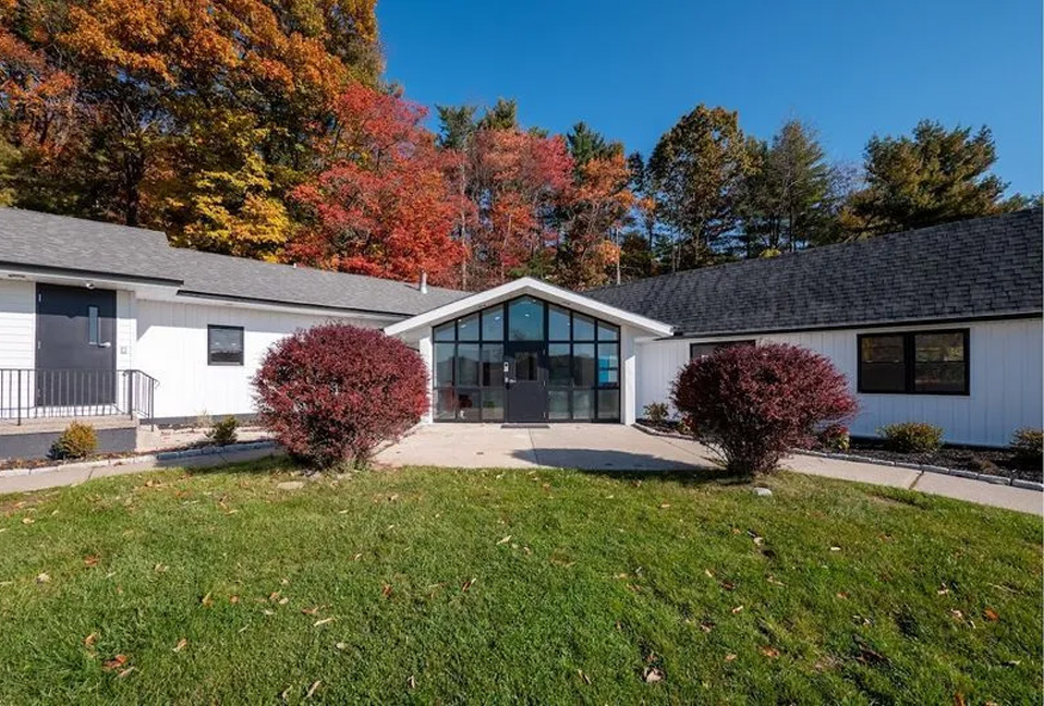 The image shows a single-story residential house with a white exterior, a black roof, and a prominent front porch. There s a covered entryway with a glass door, and the property is surrounded by a manicured lawn with some fallen leaves scattered around, indicating autumn season. The house is situated in a suburban setting with other houses visible in the background, and there are trees with fall foliage along the street.