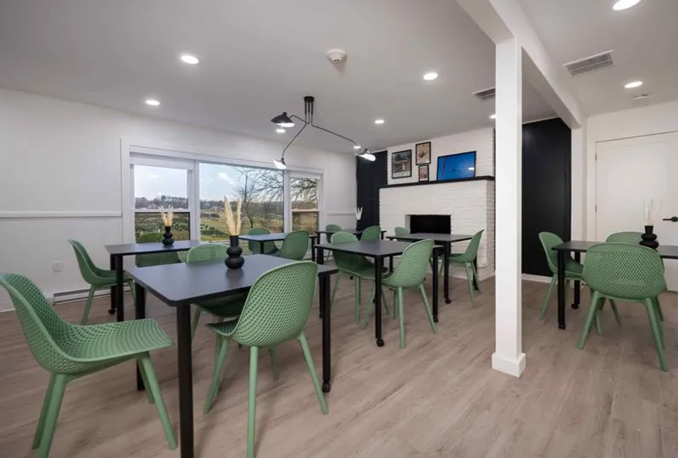 The image shows an interior space with a modern design, featuring a dining area with a table surrounded by chairs, a kitchen counter, and a view of a living room through a doorway.
