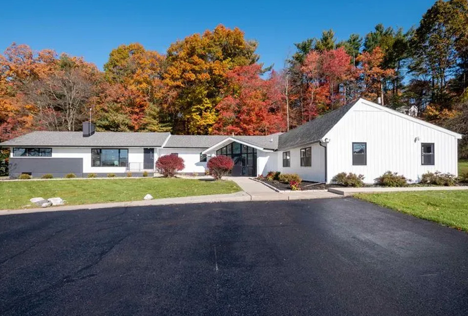 The image depicts a single-story white building with a black roof, situated on a paved driveway surrounded by green grass and trees with autumn foliage.