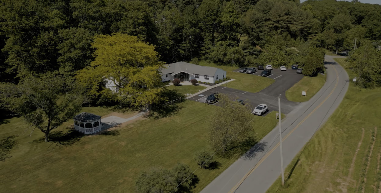 The image depicts a rural landscape with a two-story house, a parking lot with several vehicles, a curved driveway leading to the house, and a clear sky above.