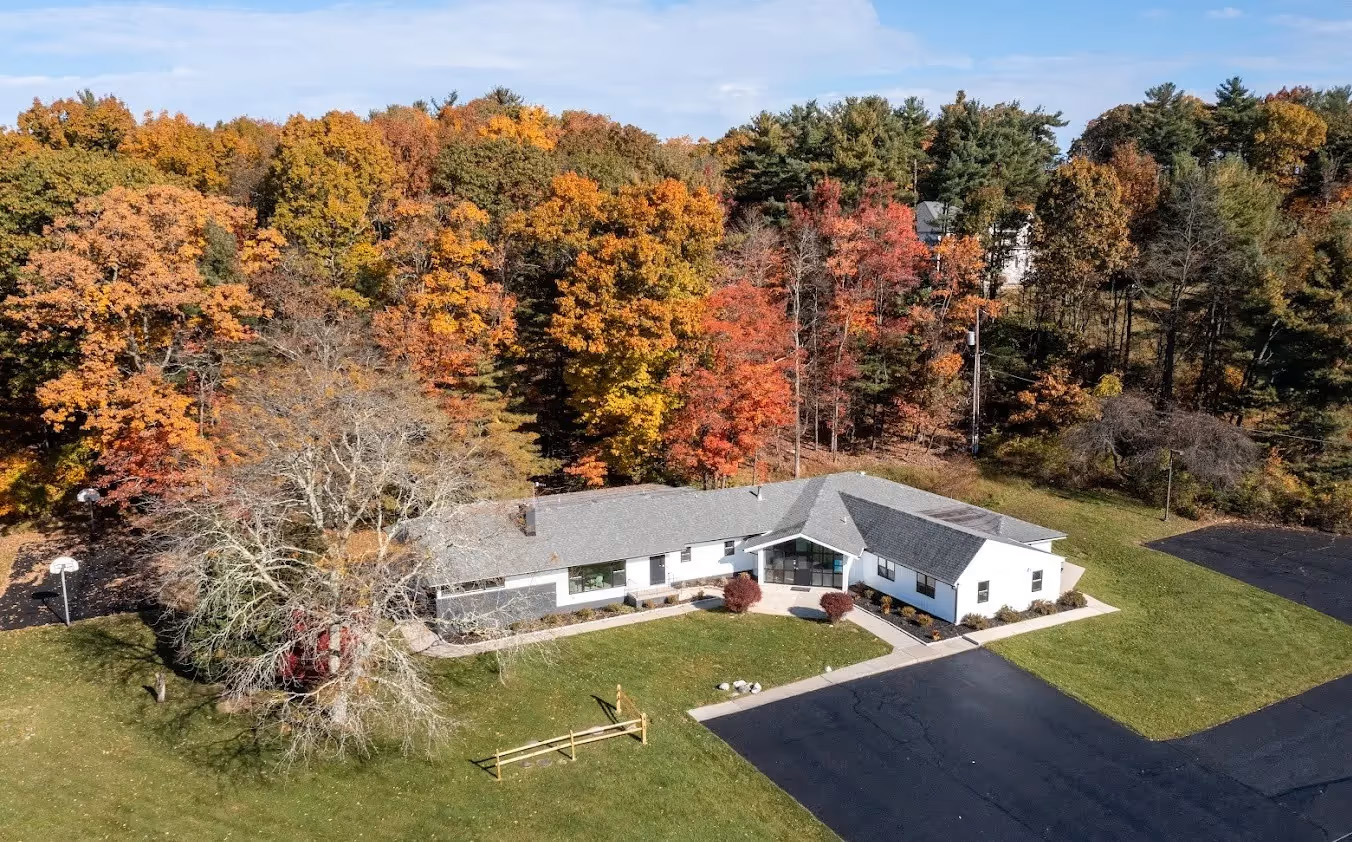 The image displays a rural residential scene with a house surrounded by trees showing autumn colors, a driveway leading up to the house, and a clear sky above.