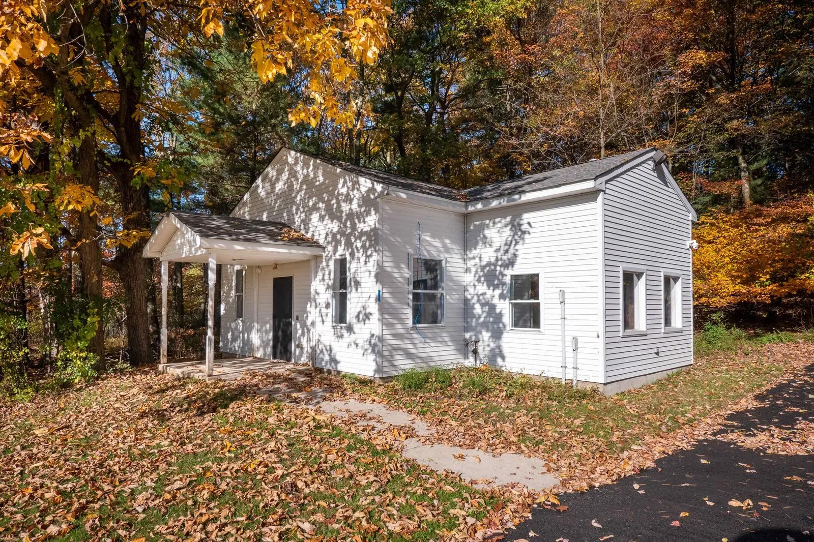 The image shows a single-story house with white siding, a gray roof, and a covered front porch during autumn, surrounded by fallen leaves.