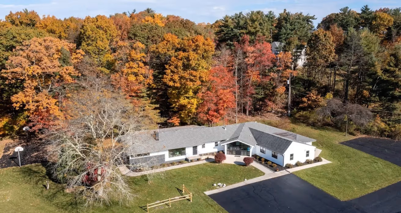 A two-story house surrounded by autumn foliage with a clear sky above and a tree standing prominently in front of the property.