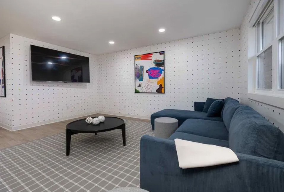 The image shows a modern living room with contemporary furniture, a patterned wall, and a large window allowing natural light into the space.