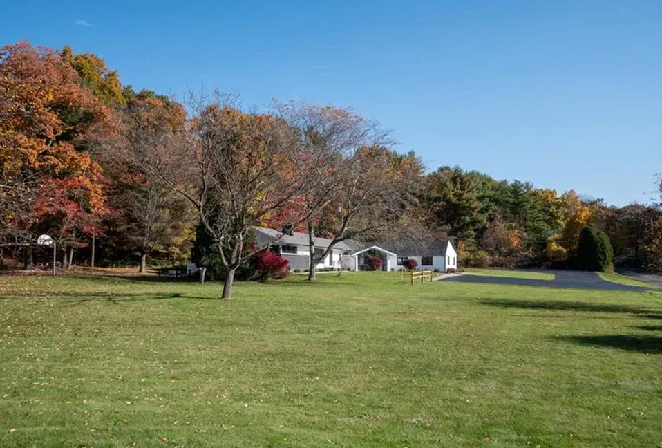 The image shows a tranquil suburban scene with a grassy yard, a house, a driveway, and trees with autumn foliage.