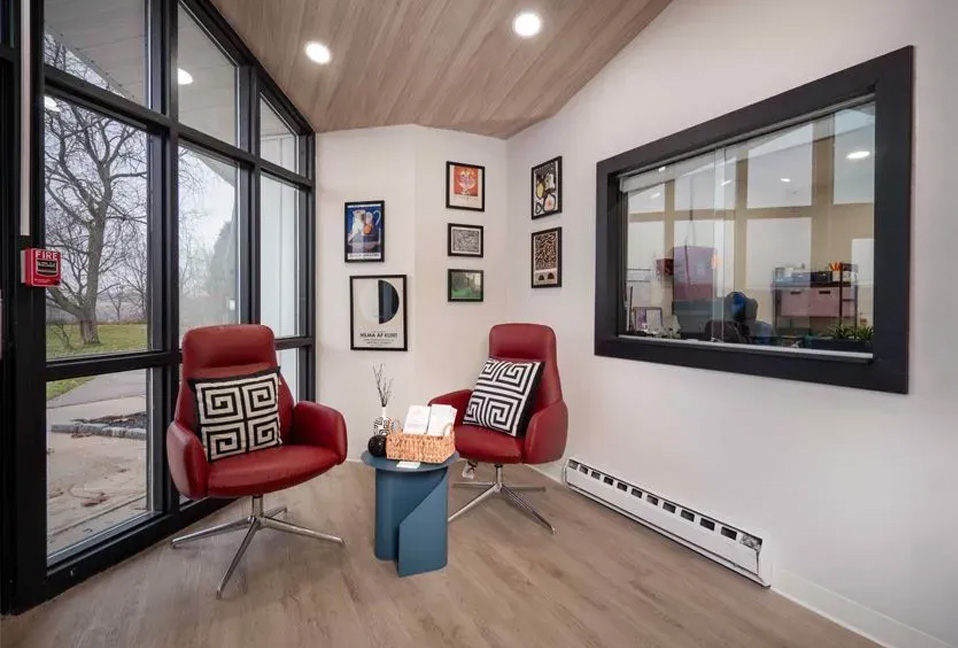 The image shows an interior space with a modern design featuring a large window, a red armchair, a desk with a chair, a wooden floor, white walls, and framed artwork on the wall.