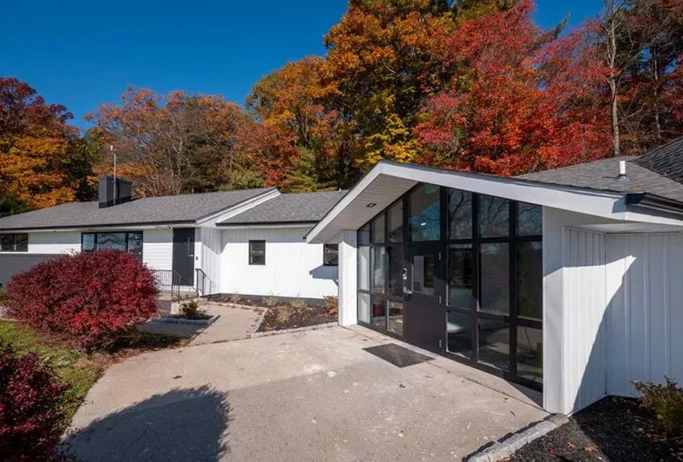 A two-story residential house with a white exterior, a prominent front porch, and a garage door, set against an autumnal landscape with red foliage.